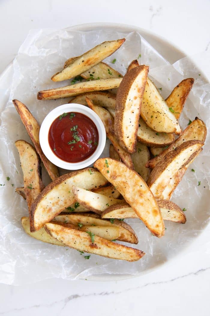 Golden brown air-fried potato wedges served on a round plate lined with parchment paper, with a small bowl of ketchup for dipping.