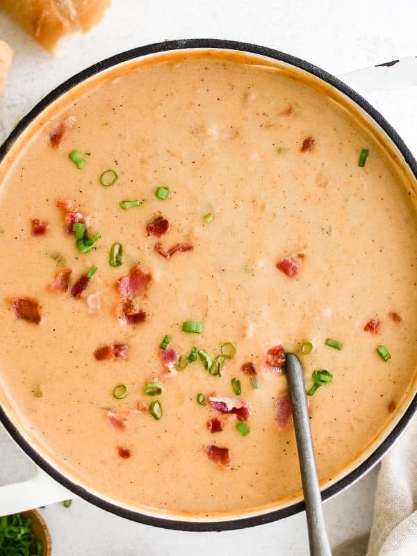 Large white pot filled with creamy orange beer cheese soup, topped with crispy bacon pieces and sliced green onions, with a spoon resting inside.