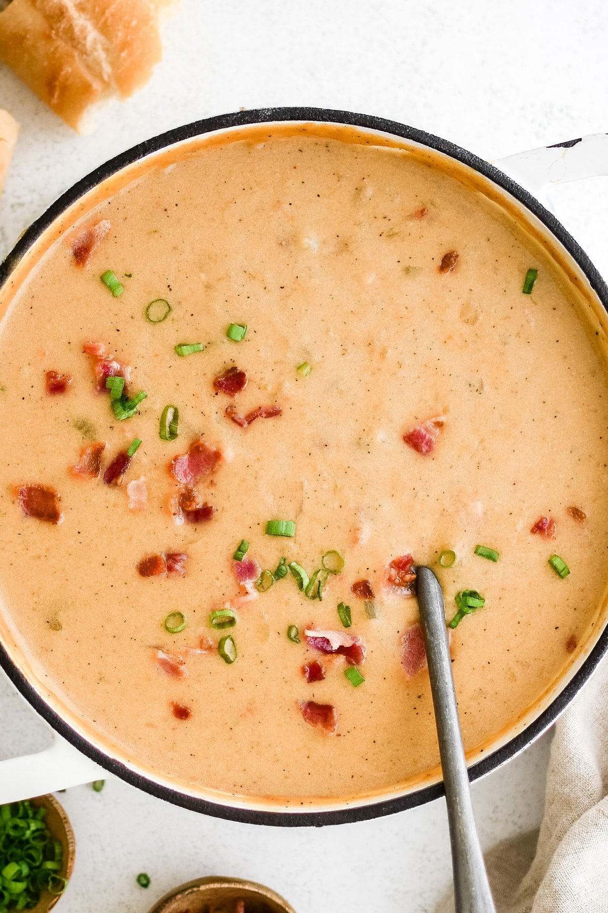 Large white pot filled with creamy orange beer cheese soup, topped with crispy bacon pieces and sliced green onions, with a spoon resting inside.