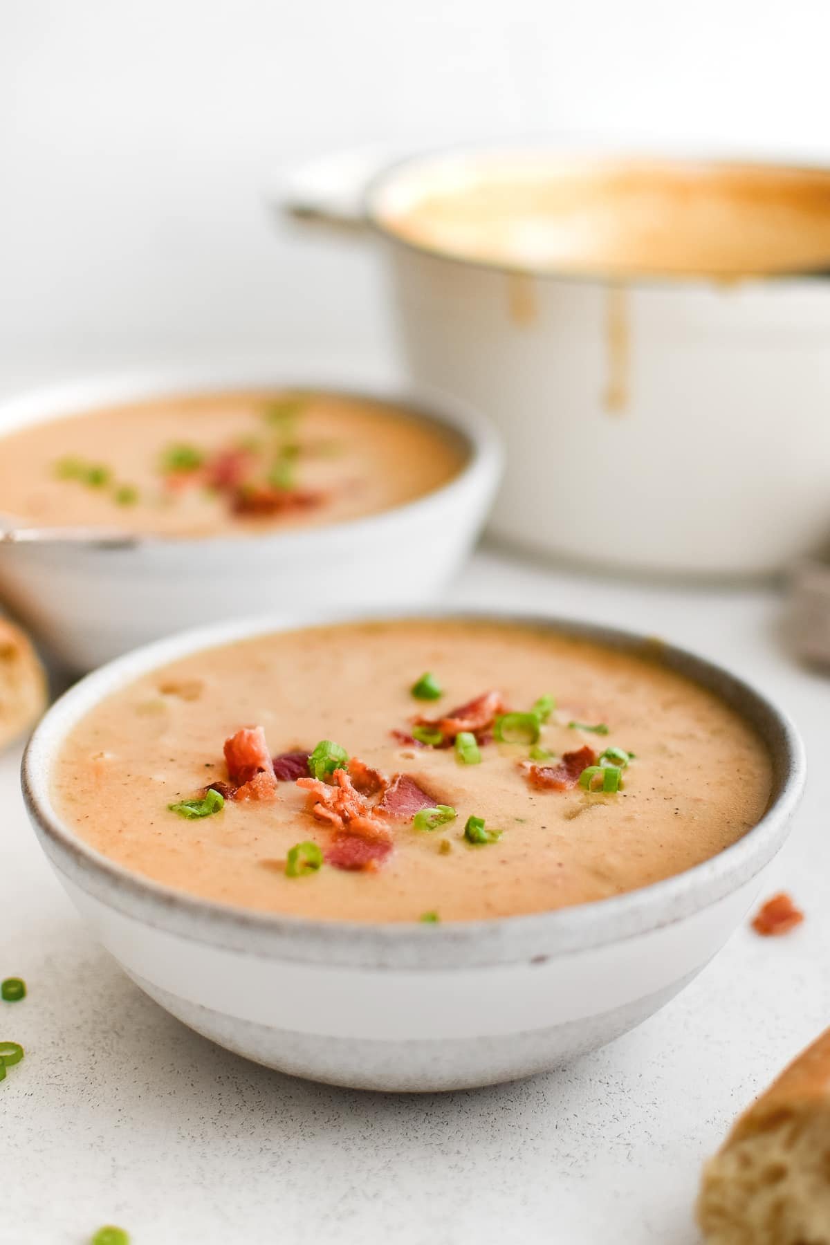 Two bowls of creamy beer cheese soup topped with bacon and green onions, served alongside a white pot of soup and slices of crusty bread.