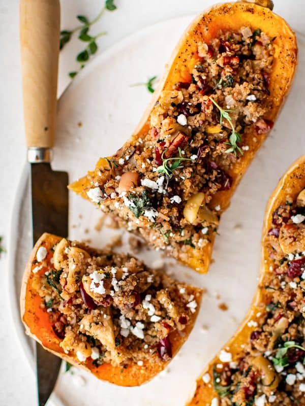 Sliced stuffed butternut squash on a white plate with a serving knife, showcasing the colorful filling of quinoa, kale, cranberries, pecans, apples, and herbs.