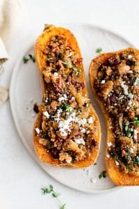 Overhead view of two stuffed butternut squash halves on a white plate, filled with quinoa, kale, apples, cranberries, pecans, and topped with fresh thyme and cheese crumbles.