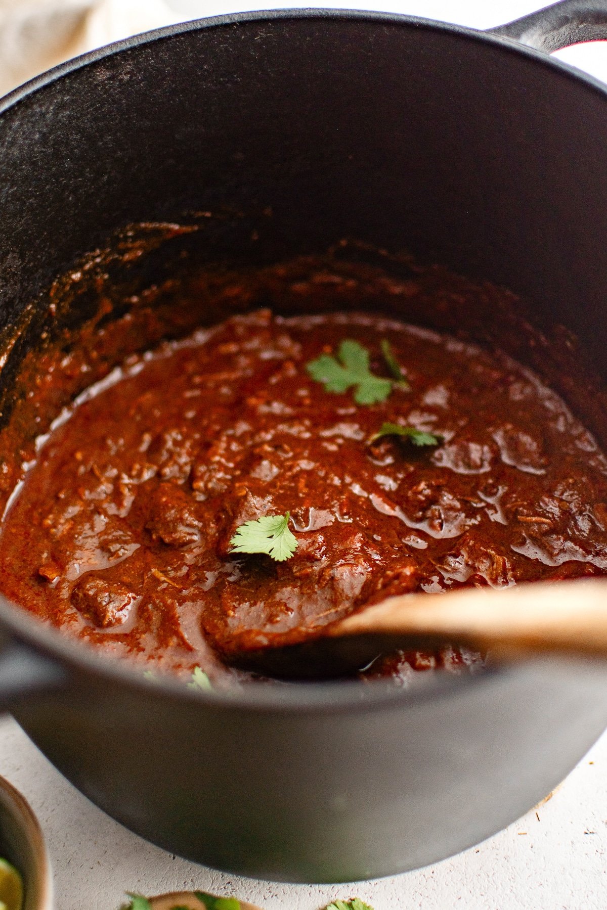 A close-up of a Dutch oven filled with thick Texas chili. The deep red sauce coats chunks of beef, with a few cilantro leaves sprinkled on top. A wooden spoon rests inside the pot, partially submerged in the chili.