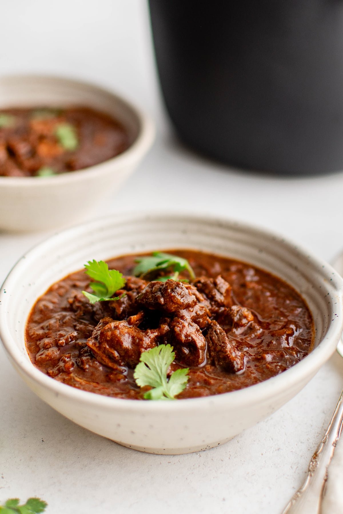 A ceramic bowl filled with Texas chili, featuring tender chunks of beef coated in a rich, smoky red sauce, topped with fresh cilantro leaves. Another bowl and the Dutch oven sit blurred in the background.