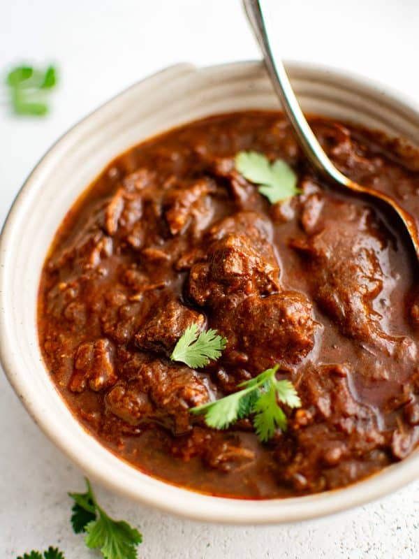 A close-up shot of a bowl of Texas chili with a spoon resting inside. Large chunks of beef sit in a thick, deep red sauce, garnished with fresh cilantro leaves scattered on top.