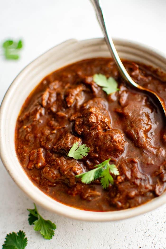 A close-up shot of a bowl of Texas chili with a spoon resting inside. Large chunks of beef sit in a thick, deep red sauce, garnished with fresh cilantro leaves scattered on top.