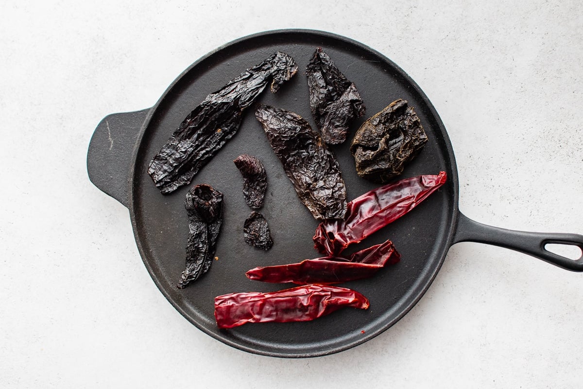 A variety of dried chili peppers, including ancho, pasilla, guajillo, and chipotle, arranged on a cast iron skillet.