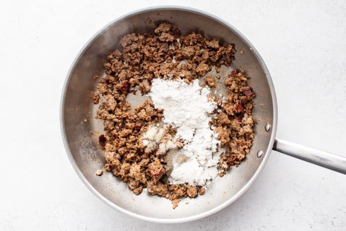 Cooked crumbled sausage in skillet topped with flour, salt, pepper, and garlic powder for making gravy.