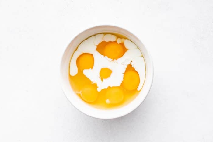 Six raw eggs and milk in a white mixing bowl before being whisked for biscuits and gravy casserole.