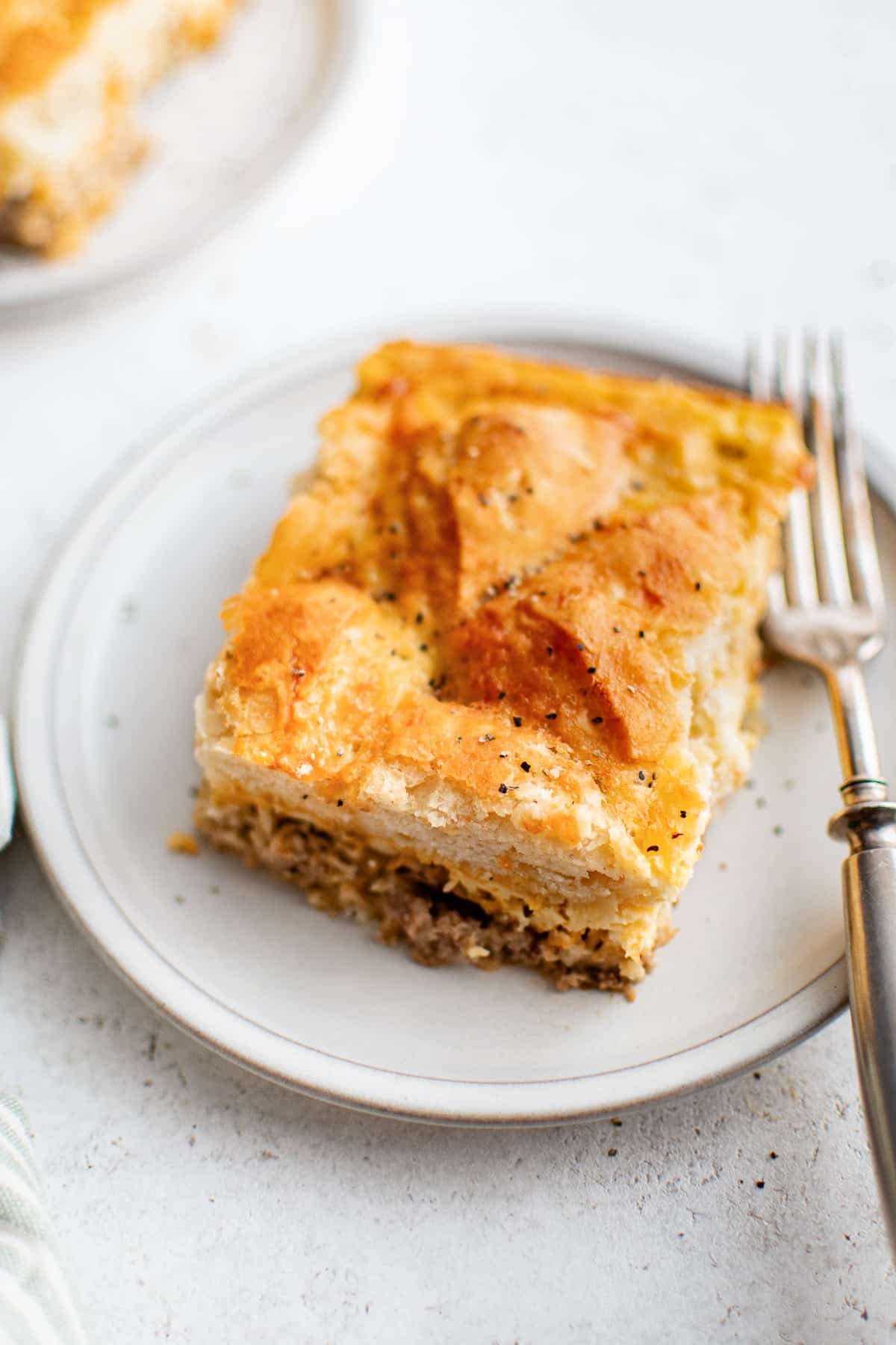 Slice of biscuits and gravy casserole on a white plate showing biscuit layers, sausage, and melted cheese.