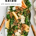 Overhead view of farro salad with apples, walnuts, arugula, and Parmesan on a white platter with lemon wedges.