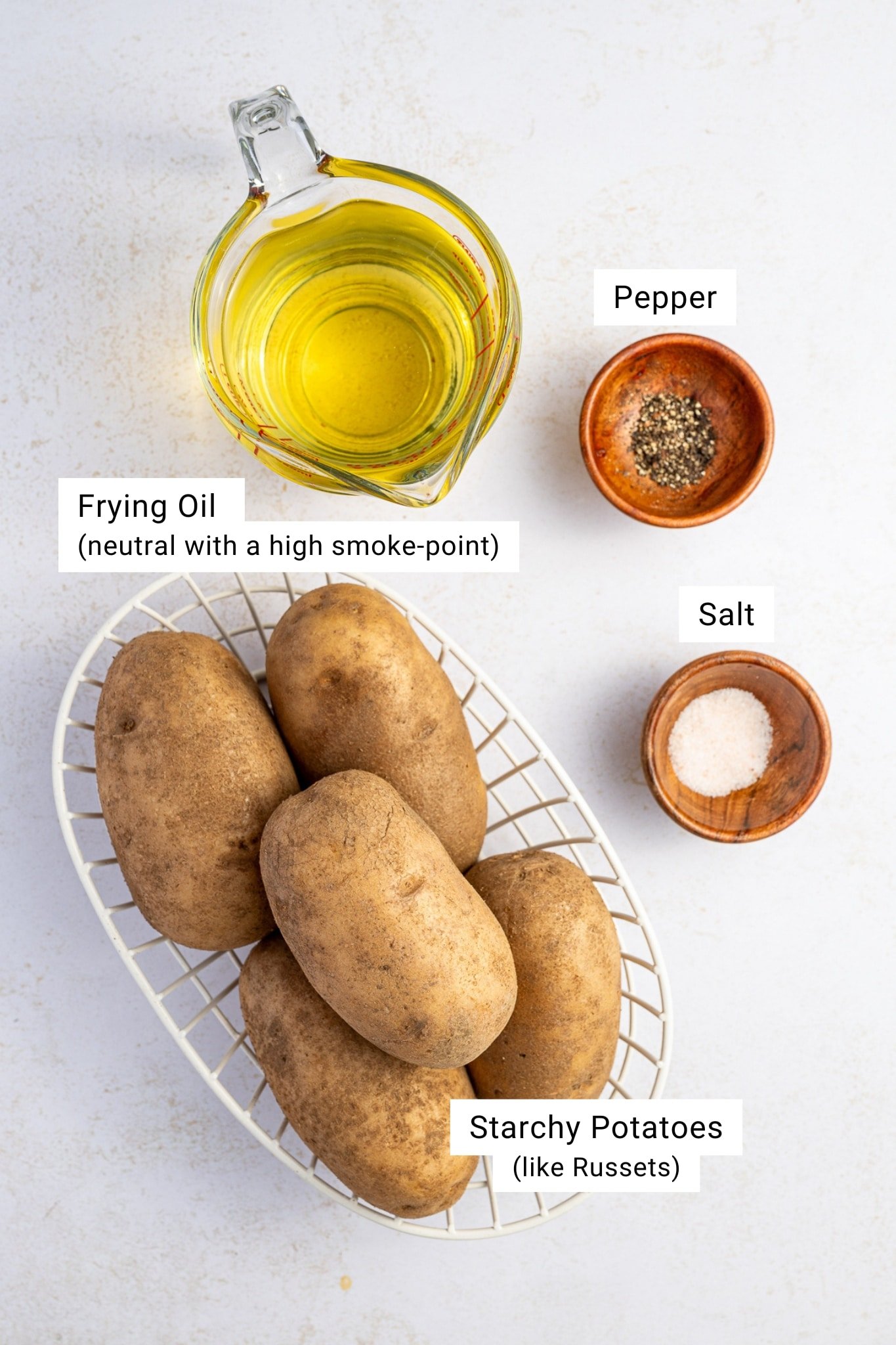 Overhead view of French fry ingredients including russet potatoes, a glass measuring cup of vegetable oil, a small bowl of salt, and a small bowl of black pepper on a white surface.