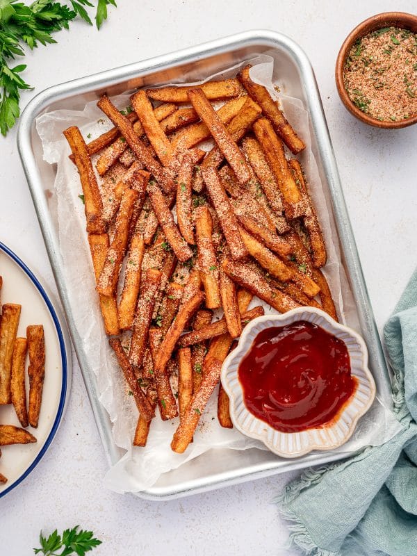 Crispy homemade French fries sprinkled with seasoning salt in a metal tray lined with parchment paper, served with a bowl of ketchup.