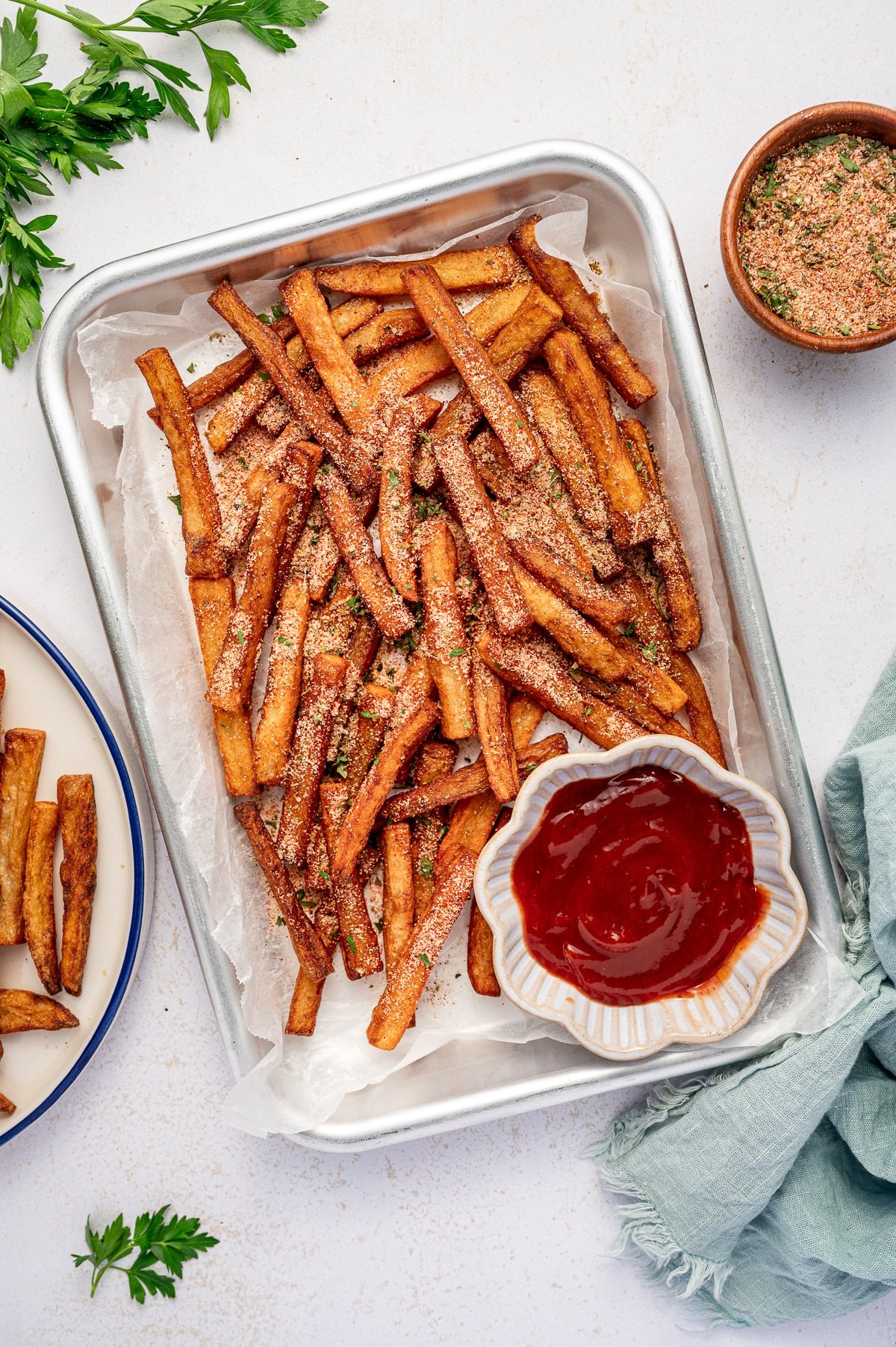 Crispy homemade French fries sprinkled with seasoning salt in a metal tray lined with parchment paper, served with a bowl of ketchup.