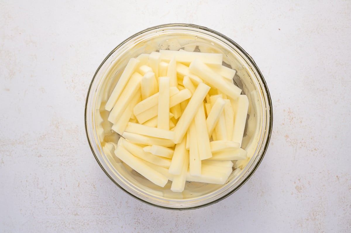 Freshly cut potato sticks are soaking in a glass bowl of cold water to remove excess starch.