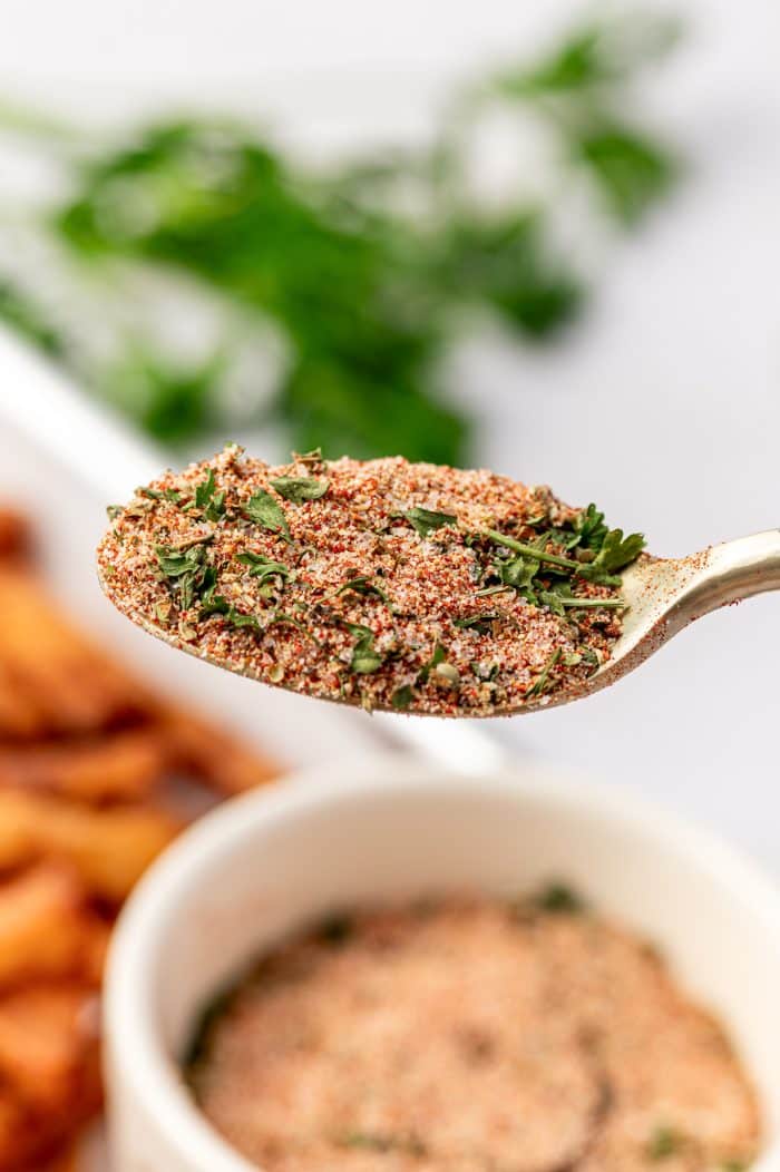 Close-up of a spoon holding homemade fry seasoning blend with visible herbs and spices, French fries blurred in the background.