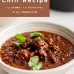 A ceramic bowl filled with smoky red Texas chili, featuring chunks of tender beef coated in rich chili sauce, garnished with cilantro leaves. A brown text overlay at the top reads โAuthentic Texas Chili Recipe โ no beans, no tomatoes, slow-simmered.โ