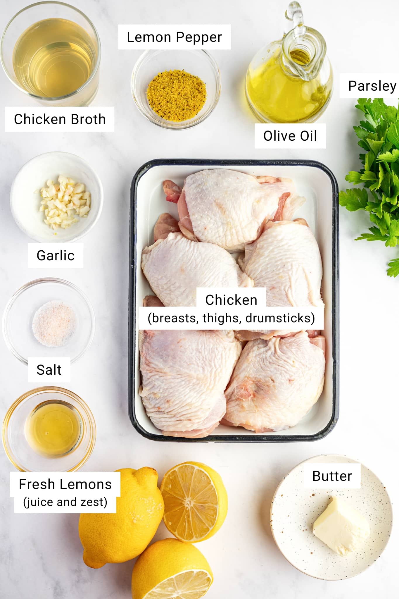 Overhead view of ingredients for baked lemon pepper chicken including raw chicken thighs, lemons, garlic, lemon pepper seasoning, olive oil, butter, and parsley.