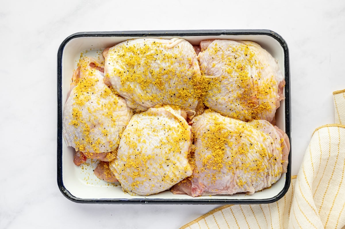 Raw chicken thighs in a white baking dish coated with lemon pepper seasoning and lemon zest.
