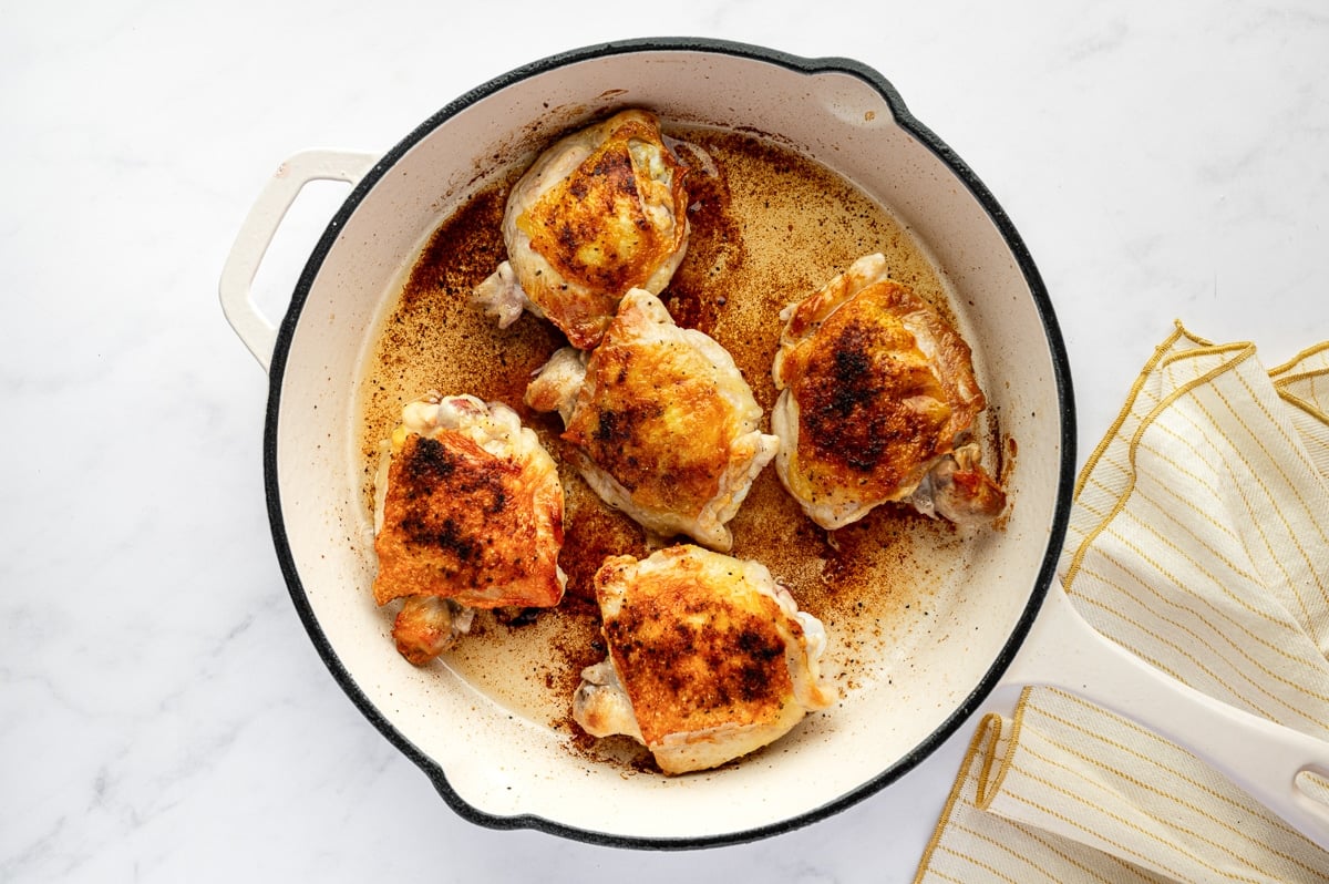 Golden seared chicken thighs in a large white oven-safe skillet after browning on the stovetop.