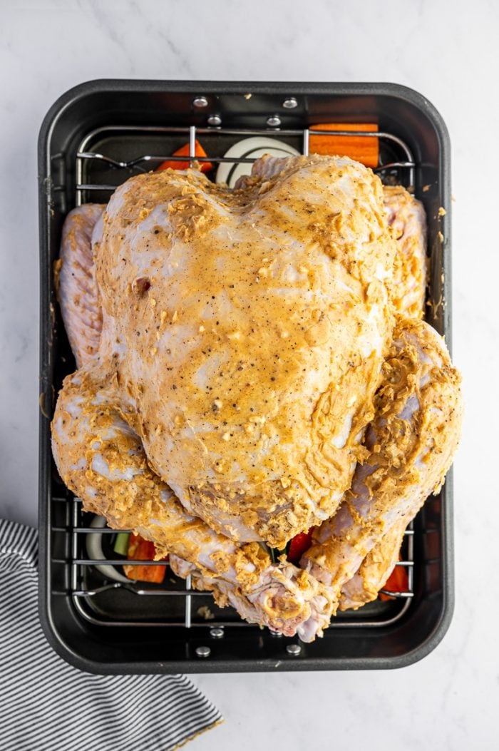 Whole turkey coated in garlic herb butter placed on a rack over vegetables before roasting.