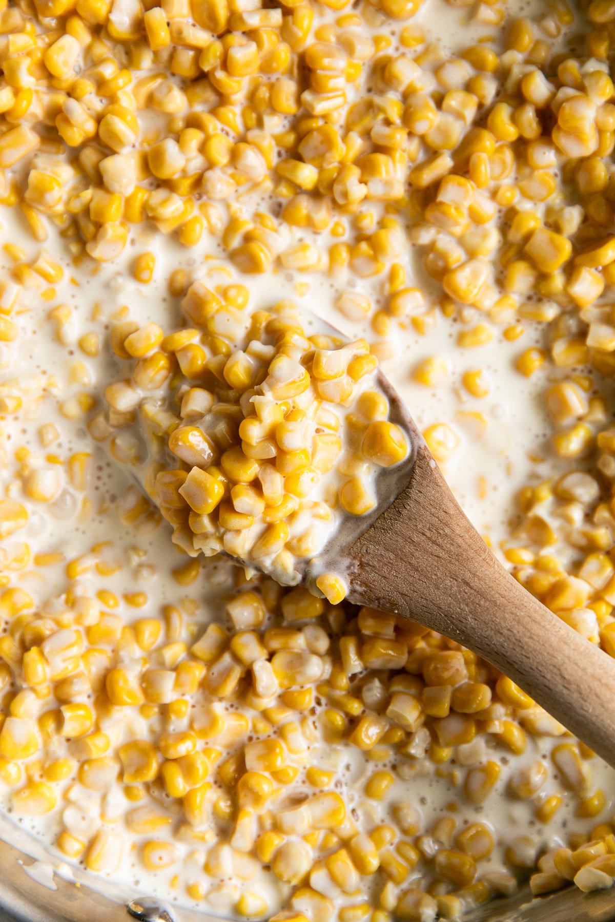 Wooden spoon stirring creamy corn in a skillet filled with rich sauce.