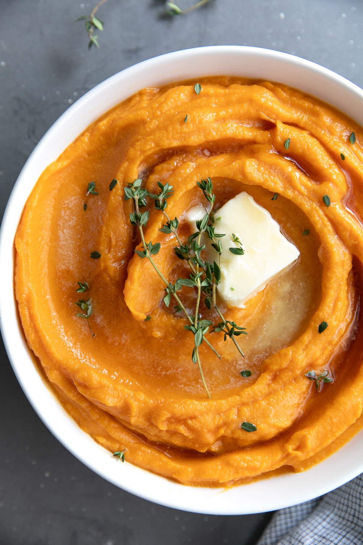 Creamy mashed sweet potatoes topped with butter and fresh thyme in a white bowl.