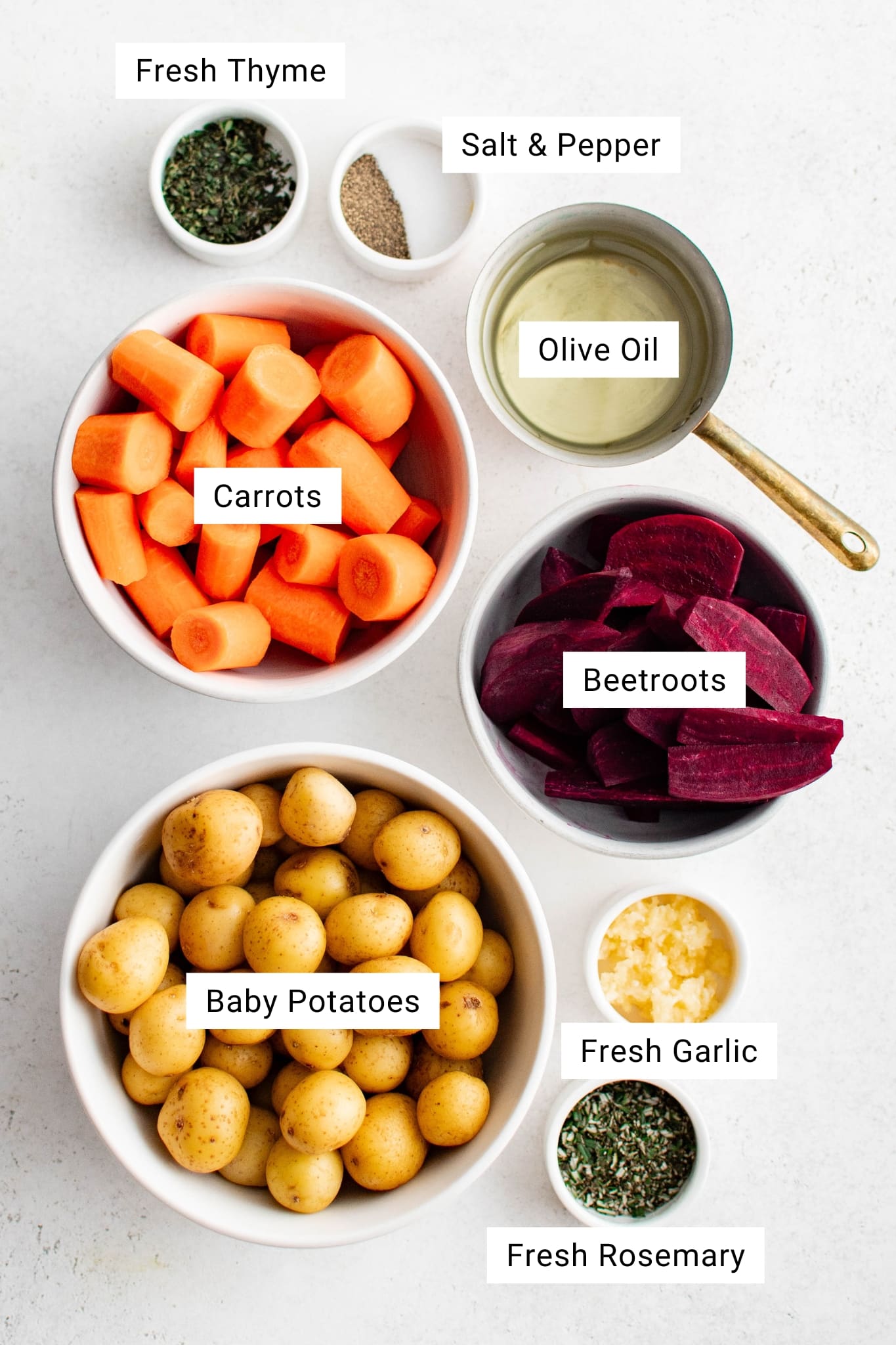 Overhead shot of labeled ingredients for roasted root vegetables including carrots, beets, baby potatoes, olive oil, garlic, rosemary, thyme, salt, and pepper.