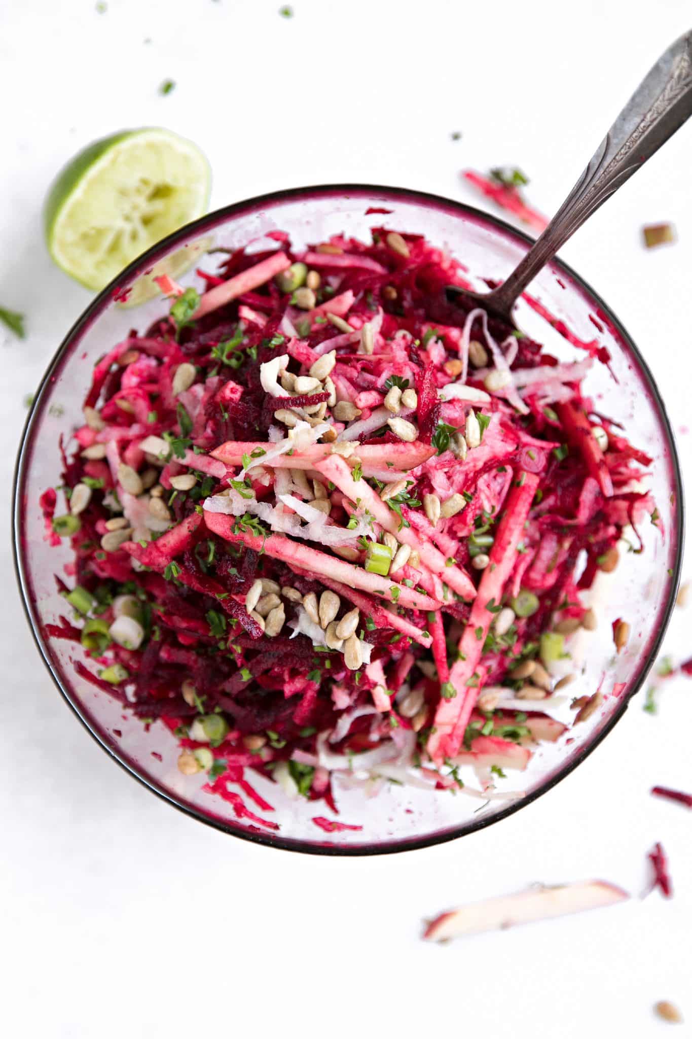 Overhead view of shredded beet and kohlrabi slaw with julienned apples, sunflower seeds, and fresh herbs in a glass bowl.