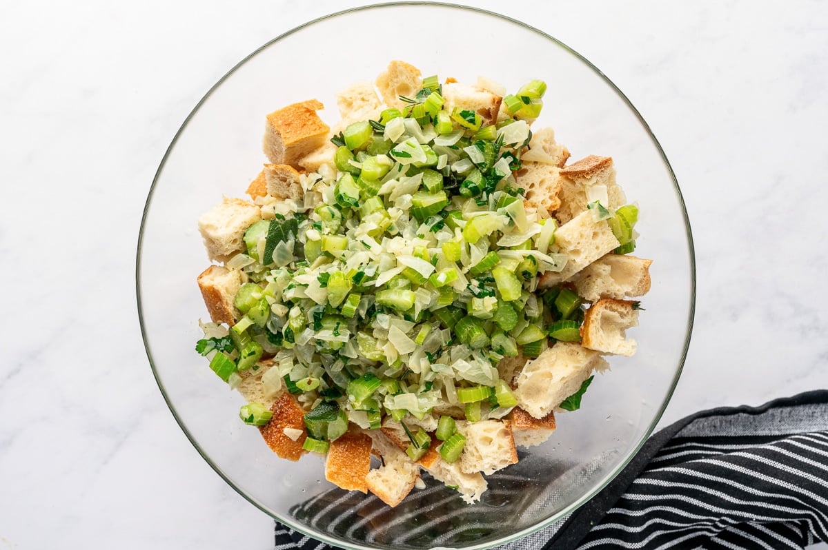 Sautéed vegetables and herbs poured over cubed bread in a large glass mixing bowl.