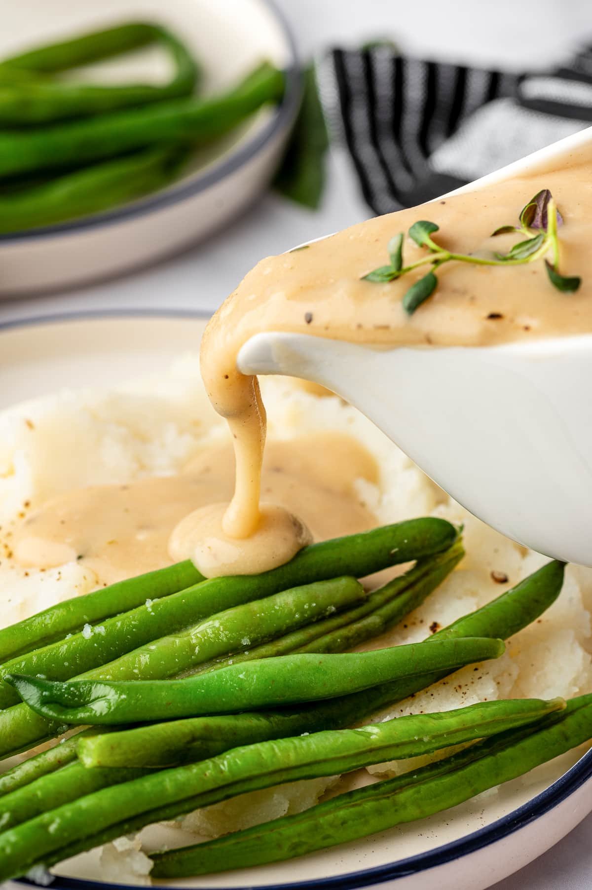 Homemade gravy being poured from a gravy boat over green beans and mashed potatoes.