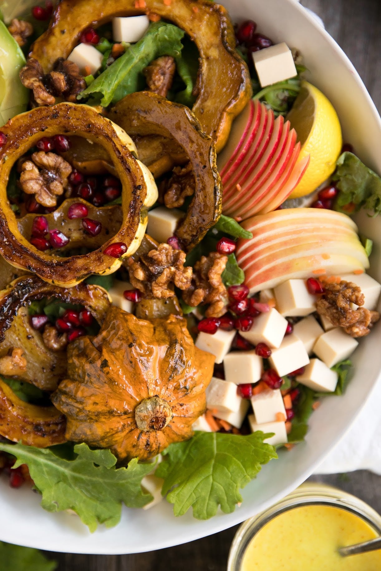 Close-up of roasted acorn squash salad with sliced apples, kale, pomegranate seeds, walnuts, and cheese cubes in a white bowl.