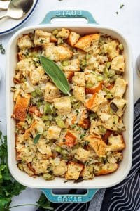 Baked Thanksgiving dressing in a blue Staub casserole dish, golden on top and garnished with fresh herbs.