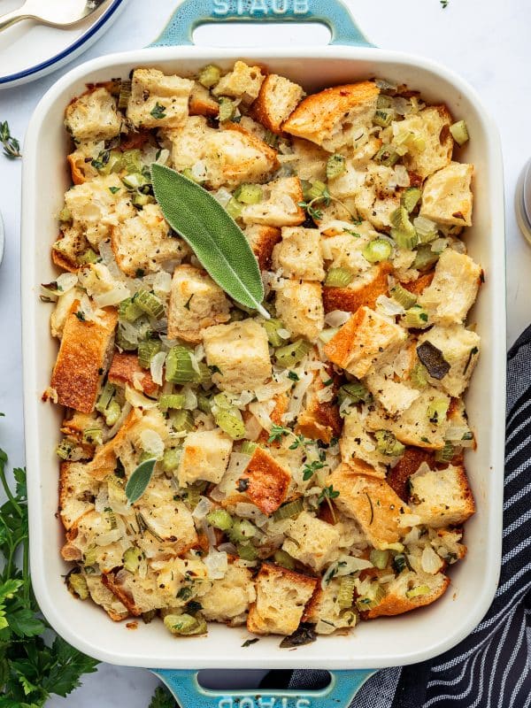 Baked Thanksgiving dressing in a blue Staub casserole dish, golden on top and garnished with fresh herbs.