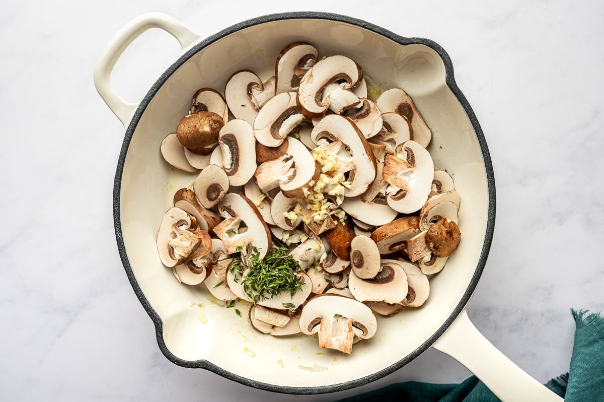 Sliced mushrooms, garlic, and fresh thyme added to sautéed shallots in a large skillet.