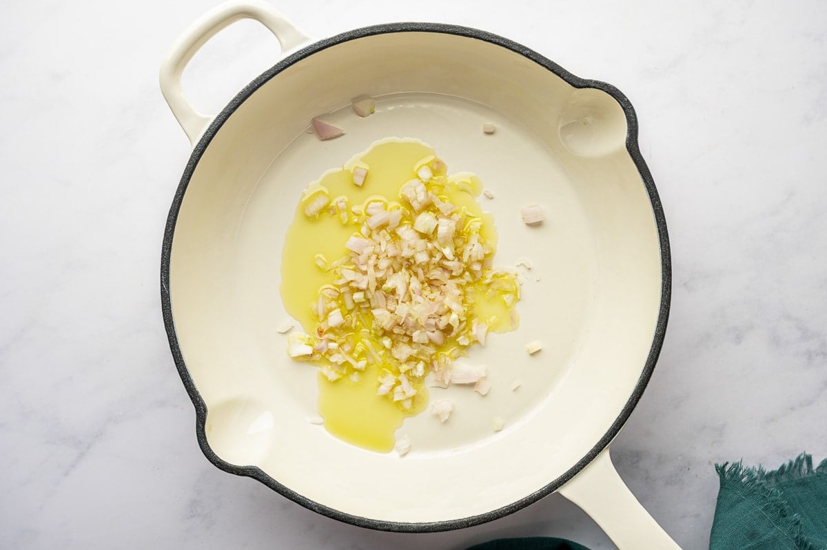 Chopped shallots sautéing in olive oil in a large cream-colored skillet.