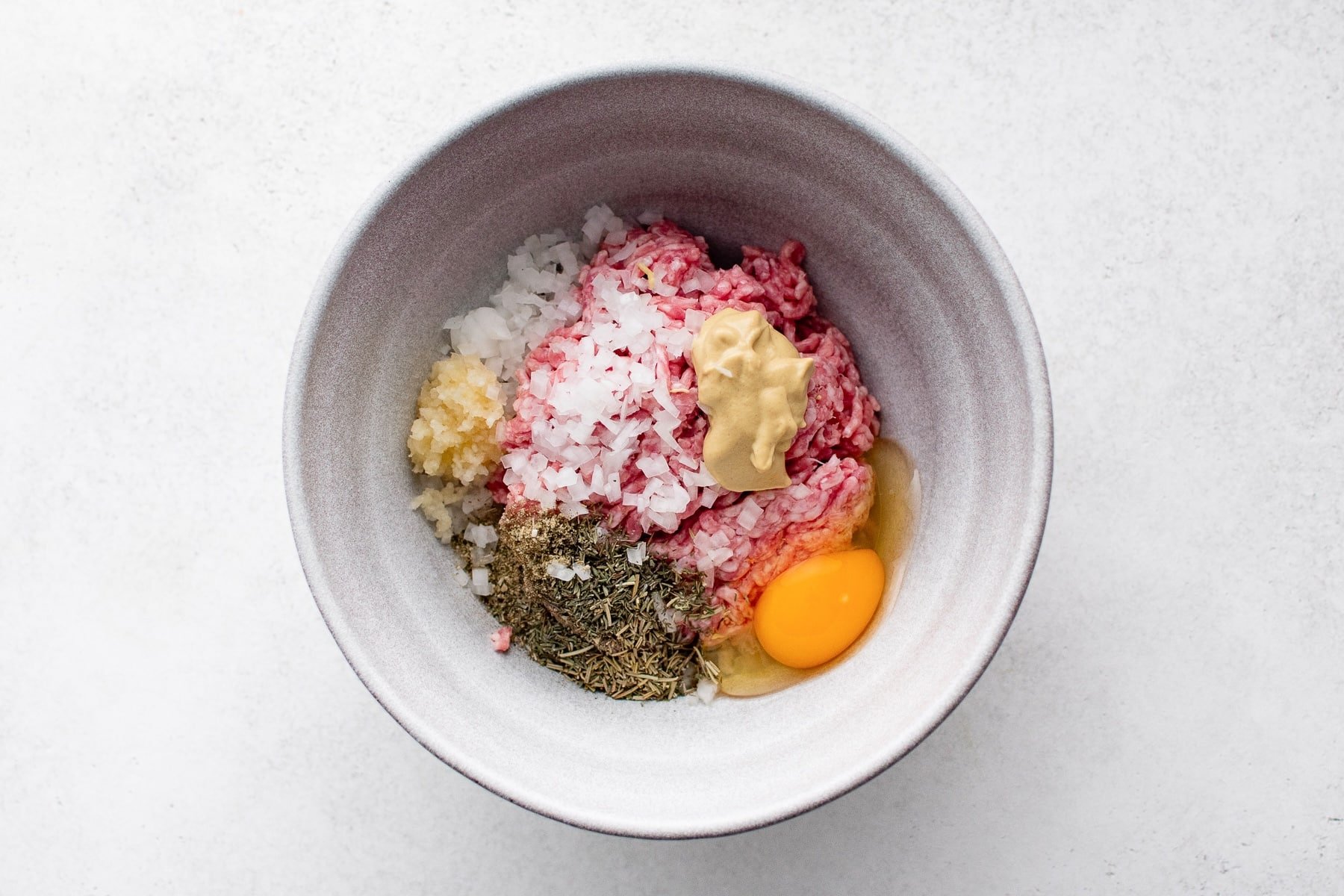 Overhead view of a gray bowl filled with raw ground pork, finely chopped onion, minced garlic, dried herbs, Dijon mustard, and a whole egg.