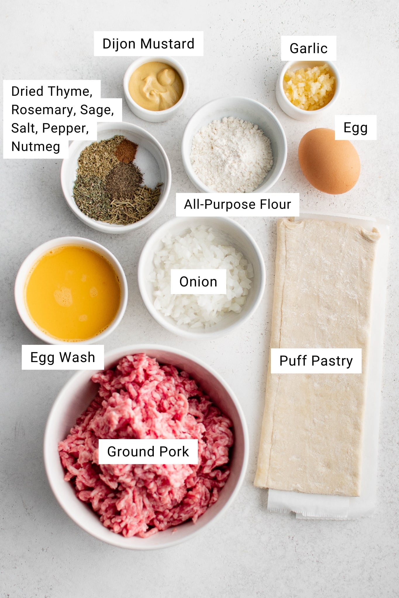 Overhead view of the ingredients for sausage rolls arranged in small bowls and labeled. Ground pork, puff pastry, chopped onion, garlic, egg, all purpose flour, dried herbs, Dijon mustard, and egg wash are displayed on a light background.