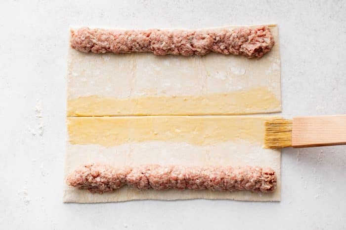 A pastry brush coats the center strip of puff pastry with egg wash while sausage filling sits along the top and bottom edges.