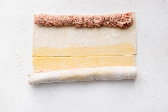 The bottom edge of the puff pastry is being rolled up tightly over the sausage mixture, creating a neat log.