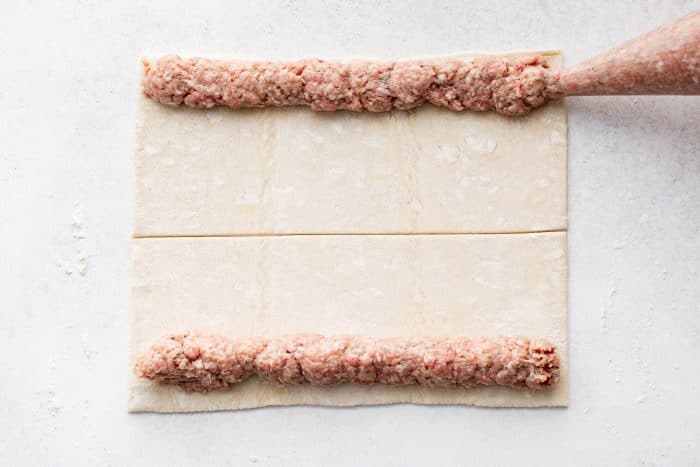 A sheet of puff pastry is laid out on a floured surface with two long, even lines of sausage filling placed along the top and bottom edges.