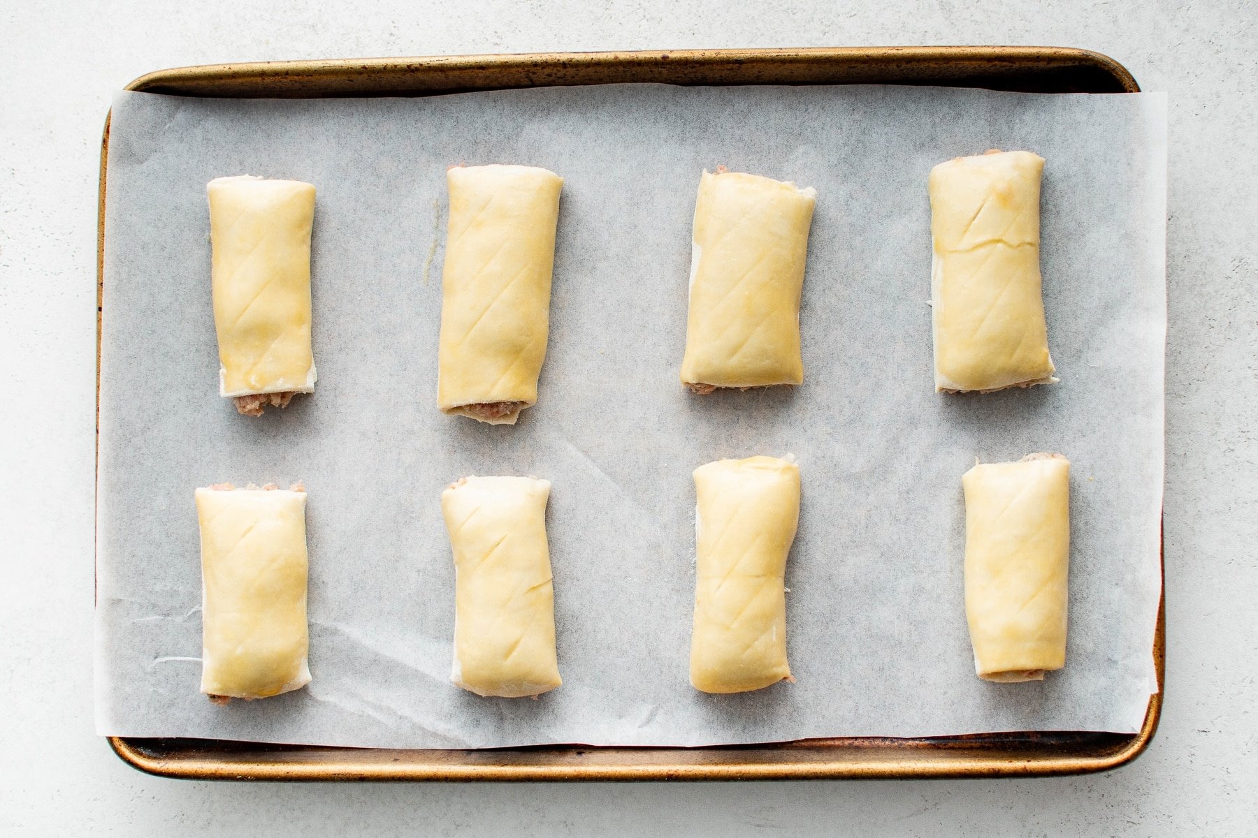 Eight evenly spaced sausage rolls sit on a parchment lined sheet pan, their puff pastry brushed with egg wash.