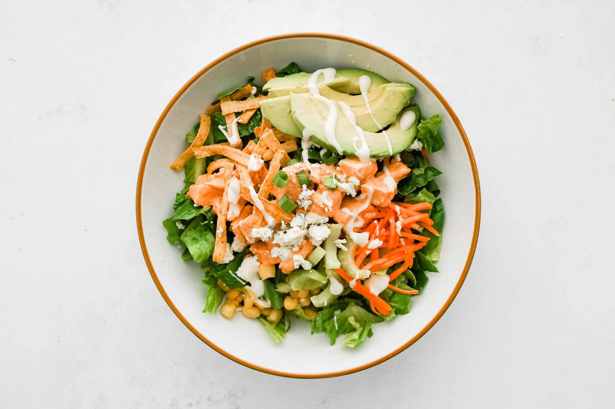 Large white salad bowl with chopped Romaine lettuce as a base topped with chicken, avocado, corn, carrots, celery, green onions, blue cheese, tortilla strips, cooked bacon, and ranch dressing.