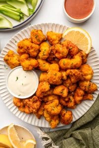 White fluted serving plate piled with crispy buffalo cauliflower bites garnished with chopped chives, served with a small white ramekin of creamy ranch dip, a lemon wedge, celery sticks on a plate in the background, and a small bowl of buffalo sauce on a light tabletop.