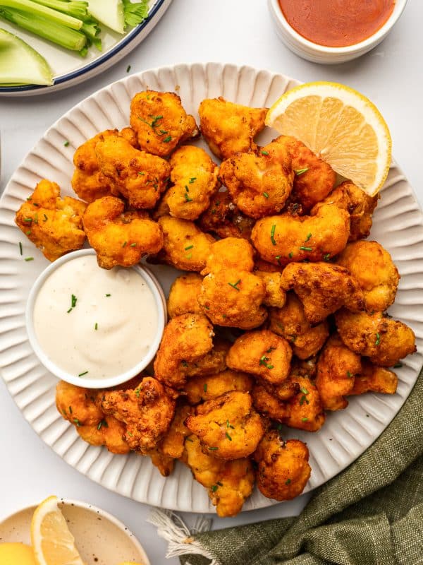 White fluted serving plate piled with crispy buffalo cauliflower bites garnished with chopped chives, served with a small white ramekin of creamy ranch dip, a lemon wedge, celery sticks on a plate in the background, and a small bowl of buffalo sauce on a light tabletop.