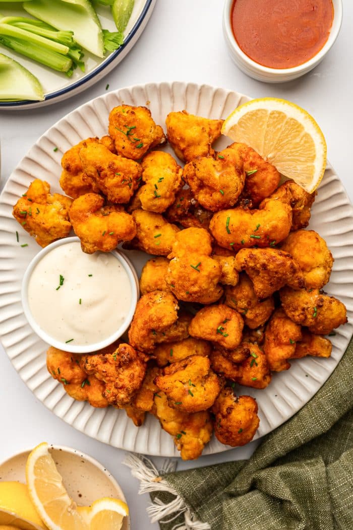 White fluted serving plate piled with crispy buffalo cauliflower bites garnished with chopped chives, served with a small white ramekin of creamy ranch dip, a lemon wedge, celery sticks on a plate in the background, and a small bowl of buffalo sauce on a light tabletop.