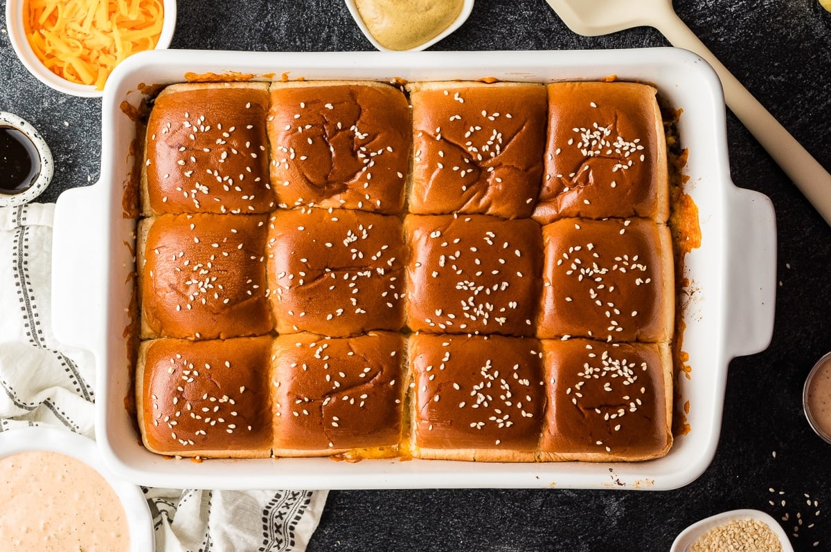 Overhead view of baked cheeseburger sliders in a white 9x13-inch baking dish, showing 12 glossy, golden-brown Hawaiian roll tops sprinkled with sesame seeds. Melted cheese and filling peek out slightly around the edges of the rolls, and small bowls of ingredients and sauce are partially visible around the pan on a dark gray textured surface.