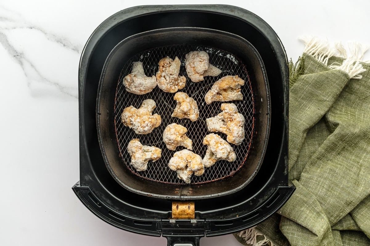 Top-down view of a black air fryer basket filled with breaded cauliflower florets coated in a pale flour mixture before cooking, sitting on a light marble countertop with a green towel on the side.
