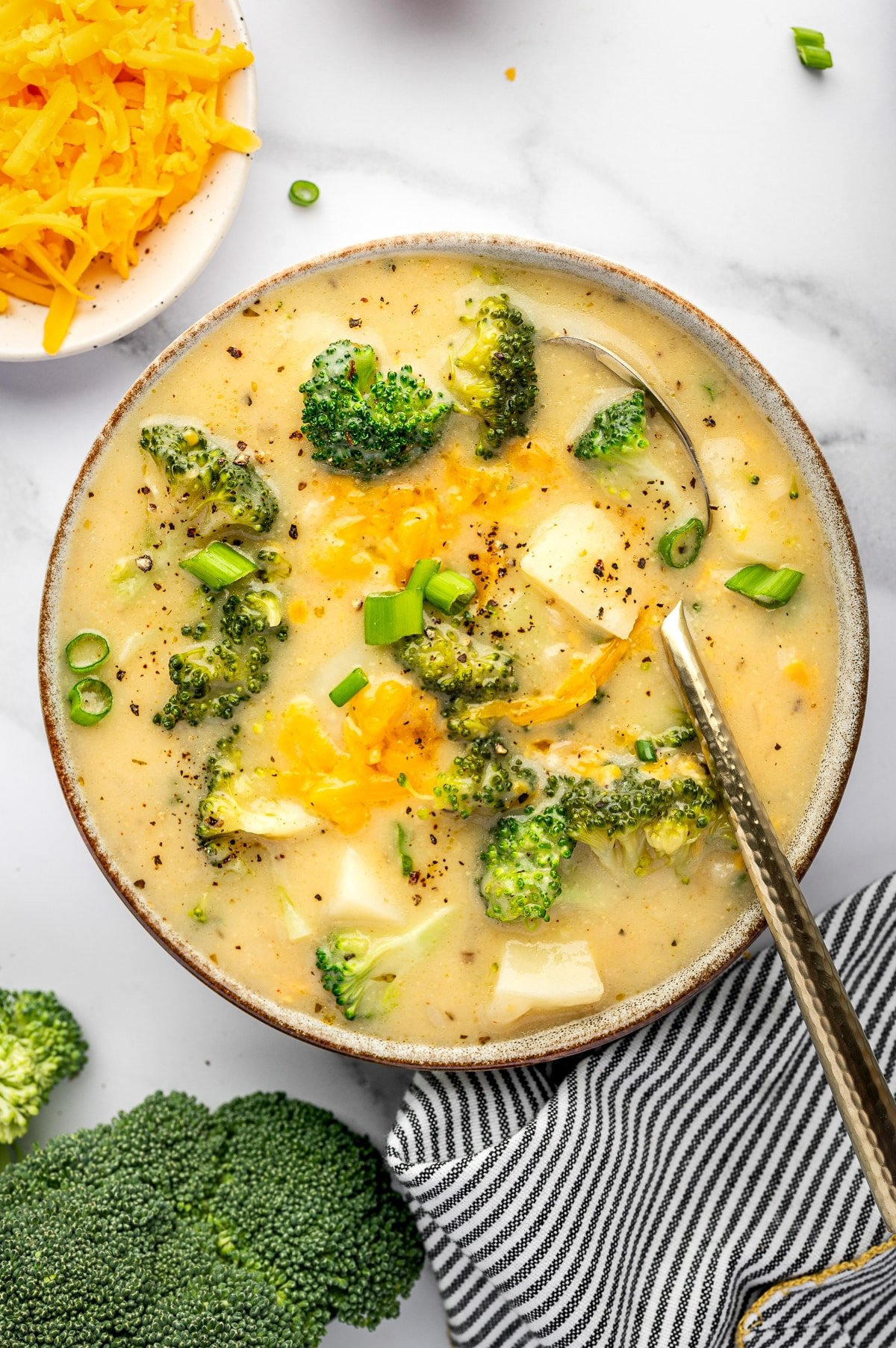 Close-up overhead view of a gray ceramic bowl with a brown rim filled with creamy broccoli potato soup on a white marble surface. Broccoli florets and potato chunks are visible, with melted cheddar cheese streaks on top, sliced green onions scattered across the surface, and cracked black pepper sprinkled throughout. A metal spoon with a textured handle rests inside the bowl on the right. A small bowl of shredded cheddar is partially visible at the top left, and a black-and-white striped towel sits at the bottom right.