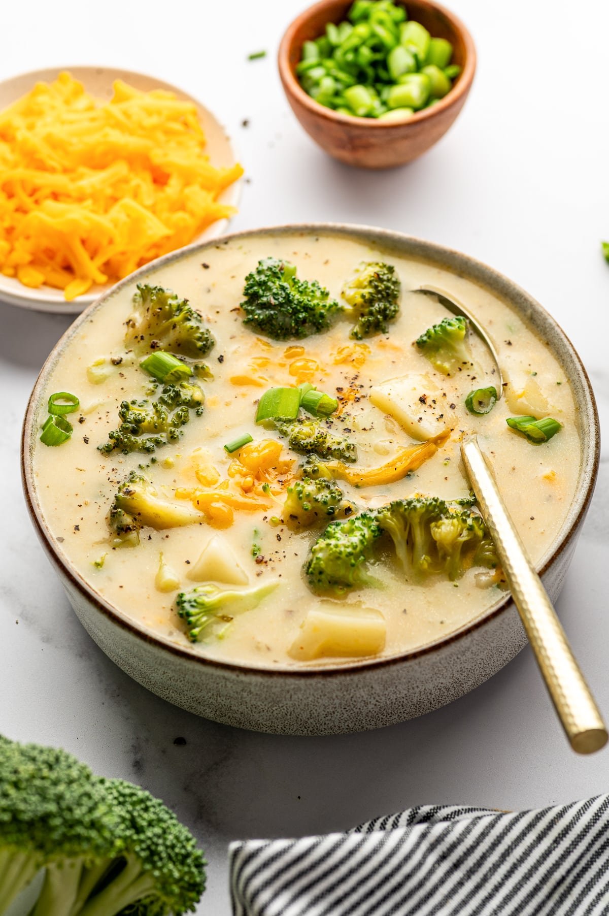Slightly wider overhead view of a gray bowl of creamy broccoli potato soup with broccoli florets, potato chunks, melted cheddar, sliced green onions, and black pepper on top. A metal spoon rests in the bowl on the right. In the background, a small wooden bowl of chopped green onions sits near the top center and a plate of shredded cheddar sits at the top left. A piece of broccoli is visible at the bottom left, and a black-and-white striped towel is draped at the bottom right on a white marble countertop.